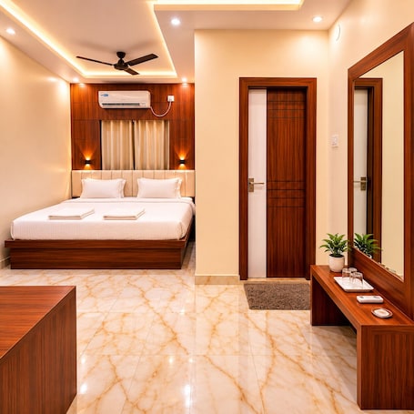 A hotel room with a bed, a ceiling fan, an air conditioning unit, a wooden door, a mirror, and a small plant on a side table.. Hotel Priyadarsini