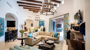 Amplia sala de estar con un sofá seccional grande, una mesa de centro y un candelabro. Se observa un comedor con mesa y sillas, y una cocina al fondo.