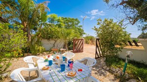 An outdoor dining area with a table set for two, surrounded by white plastic chairs, under a clear sky.