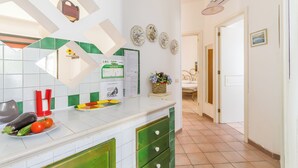A kitchen with a white countertop, green cabinets, and a tiled backsplash. There are plates with vegetables on the counter, a basket with flowers, and a wall with framed pictures.