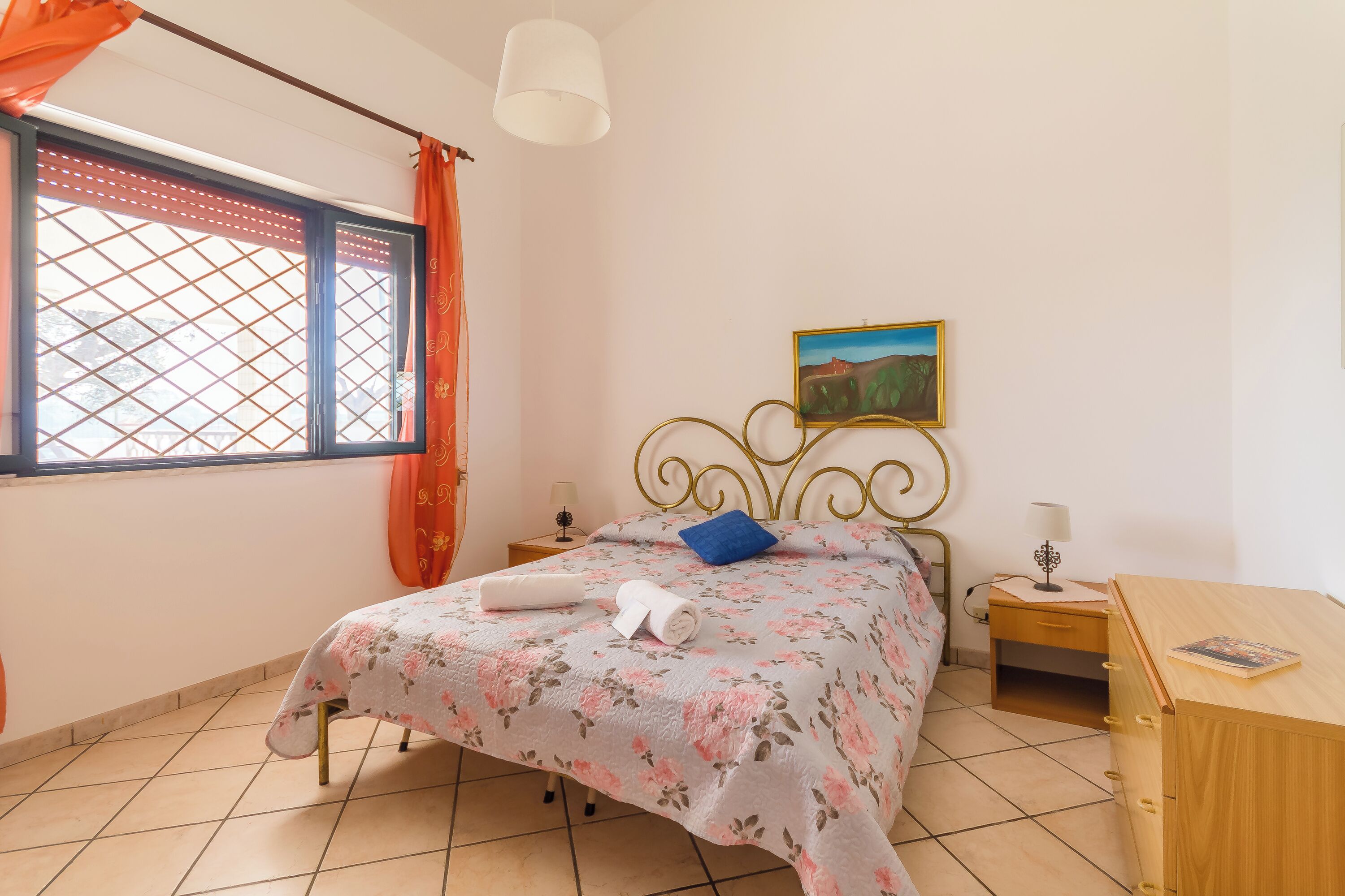 Een slaapkamer met een bed, een nachtkastje, een houten dressoir, een raam met gordijnen en een schilderij aan de muur.