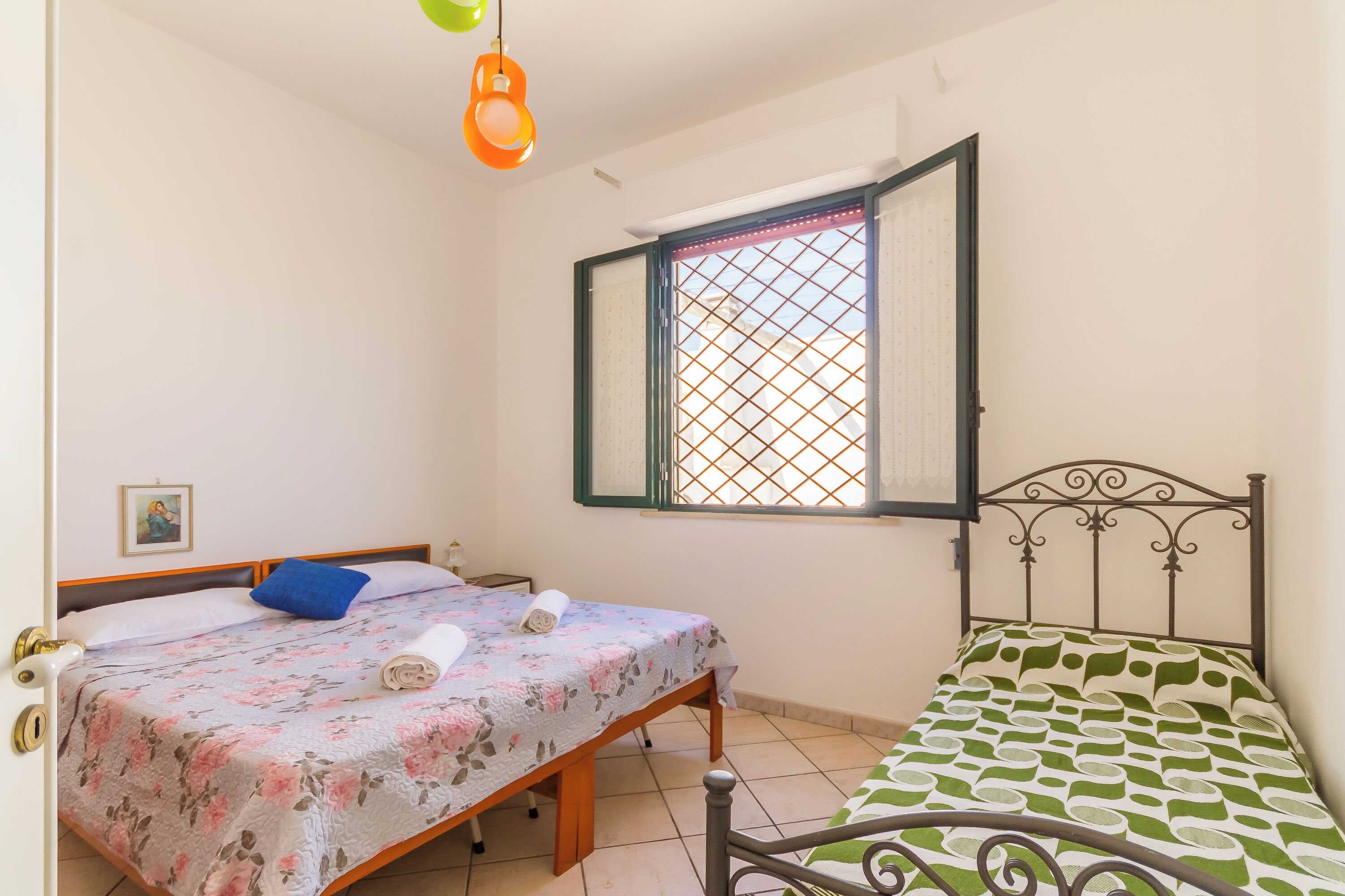 A bedroom with two beds, a window with a metal grid, and a hanging light fixture.