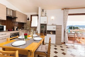 A modern kitchen with wooden cabinets, a dining table set for two, and a view of the sea through a balcony. - La Lampara House (Arzachena)