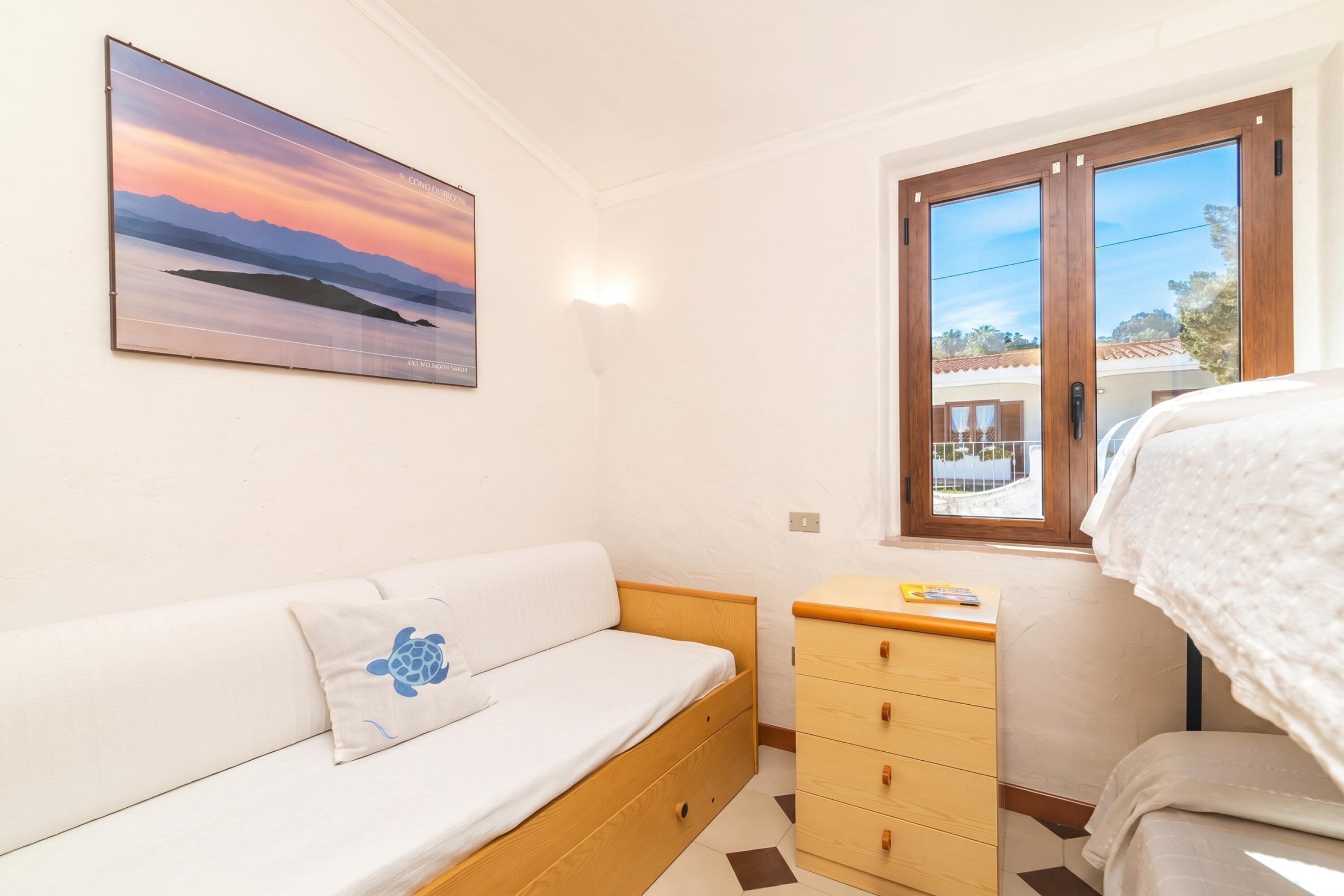 A bedroom with a bed, a wooden dresser, a sofa, a wall-mounted picture, and a window with a view.