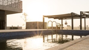 Une piscine avec un bord carrelé, une structure en forme de pergola et un bâtiment en béton en arrière-plan.