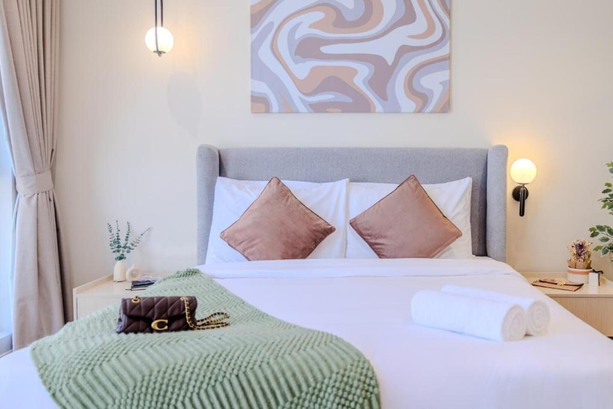 Una cama bien hecha con almohadas, una manta verde y una toalla enrollada. Encima de la cama hay una obra de arte en la pared, una lámpara de noche y una pequeña planta en la mesita.
