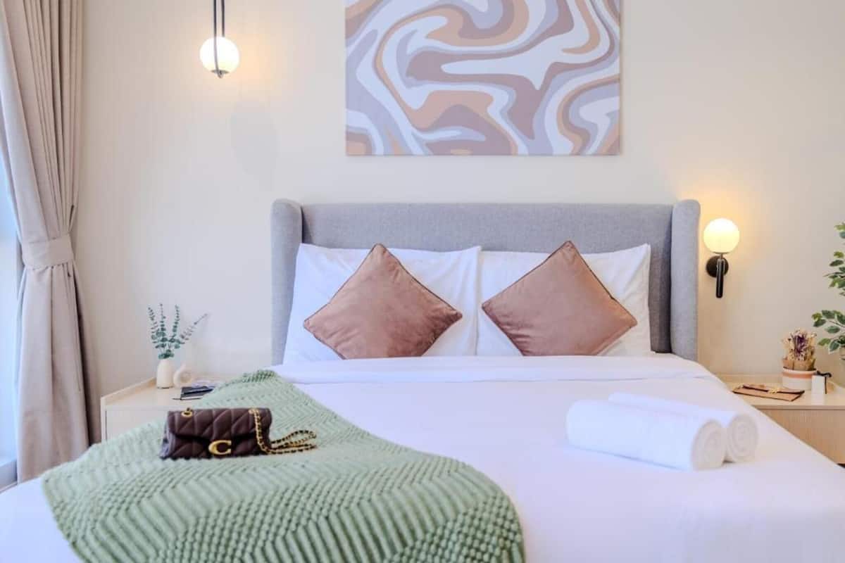 Ein ordentlich bezogenes Bett mit Kissen, einer grünen Decke und einem aufgerollten Handtuch. Darüber hängt ein Wandbild, neben dem Bett steht eine Nachttischlampe und ein kleiner Topf mit Pflanze auf dem Nachttisch.