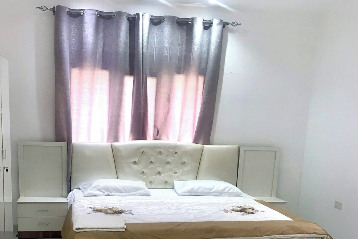 A bedroom with a large bed, a bedside table, a nightstand, and a window with curtains.