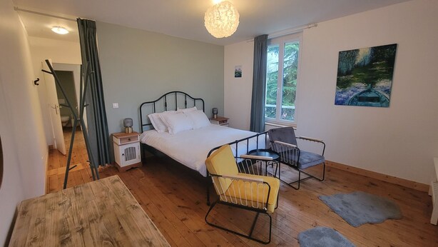 Quarto com cama, duas cadeiras, mesa, pintura e janela com vista para árvores.