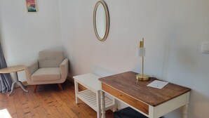 Quarto pequeno e bem arrumado, com uma secretária de madeira, uma cadeira, uma mesinha e um abajur.