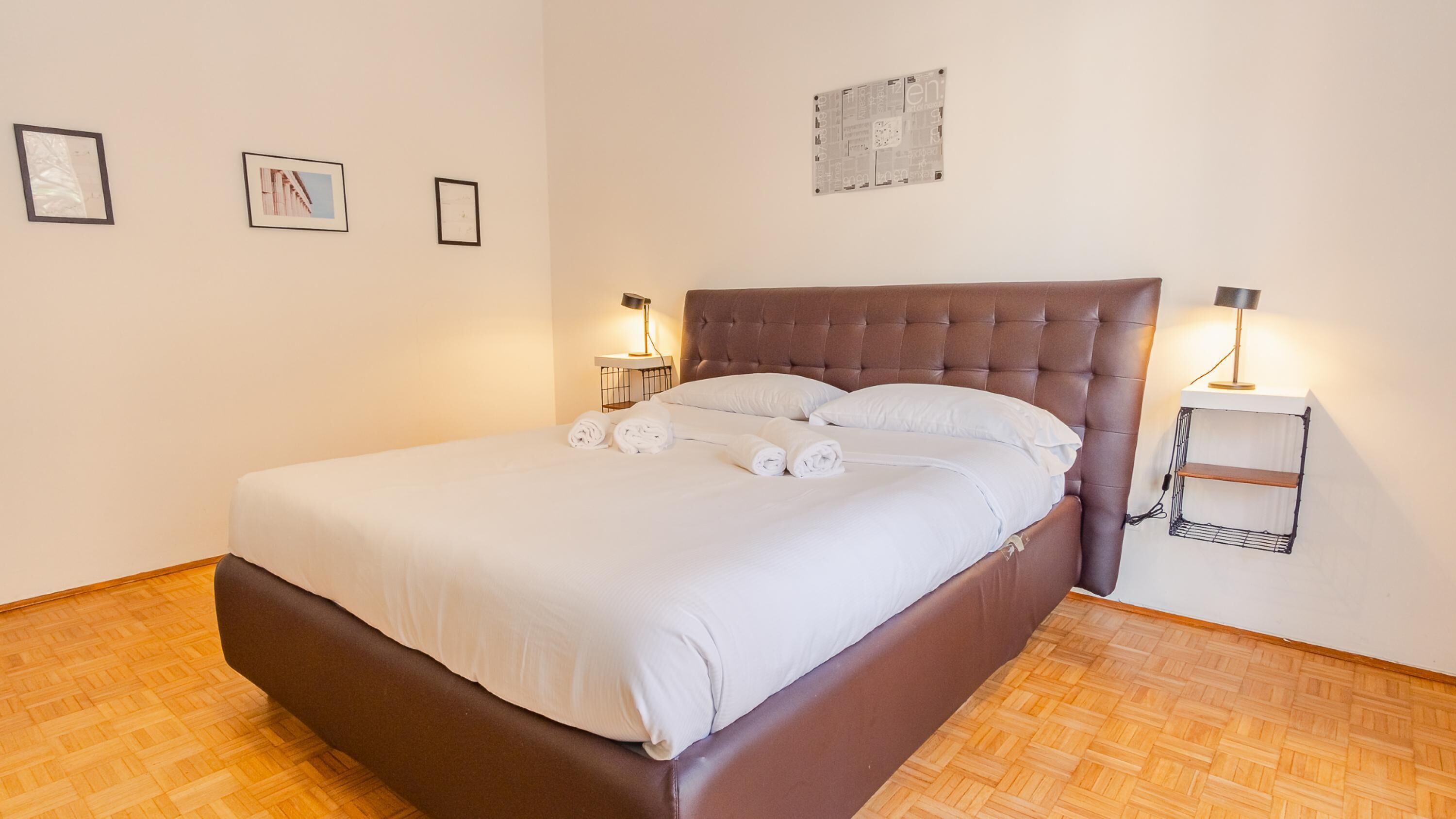 A neatly made bed with white linens and rolled towels, a brown upholstered headboard, and two bedside tables with lamps.