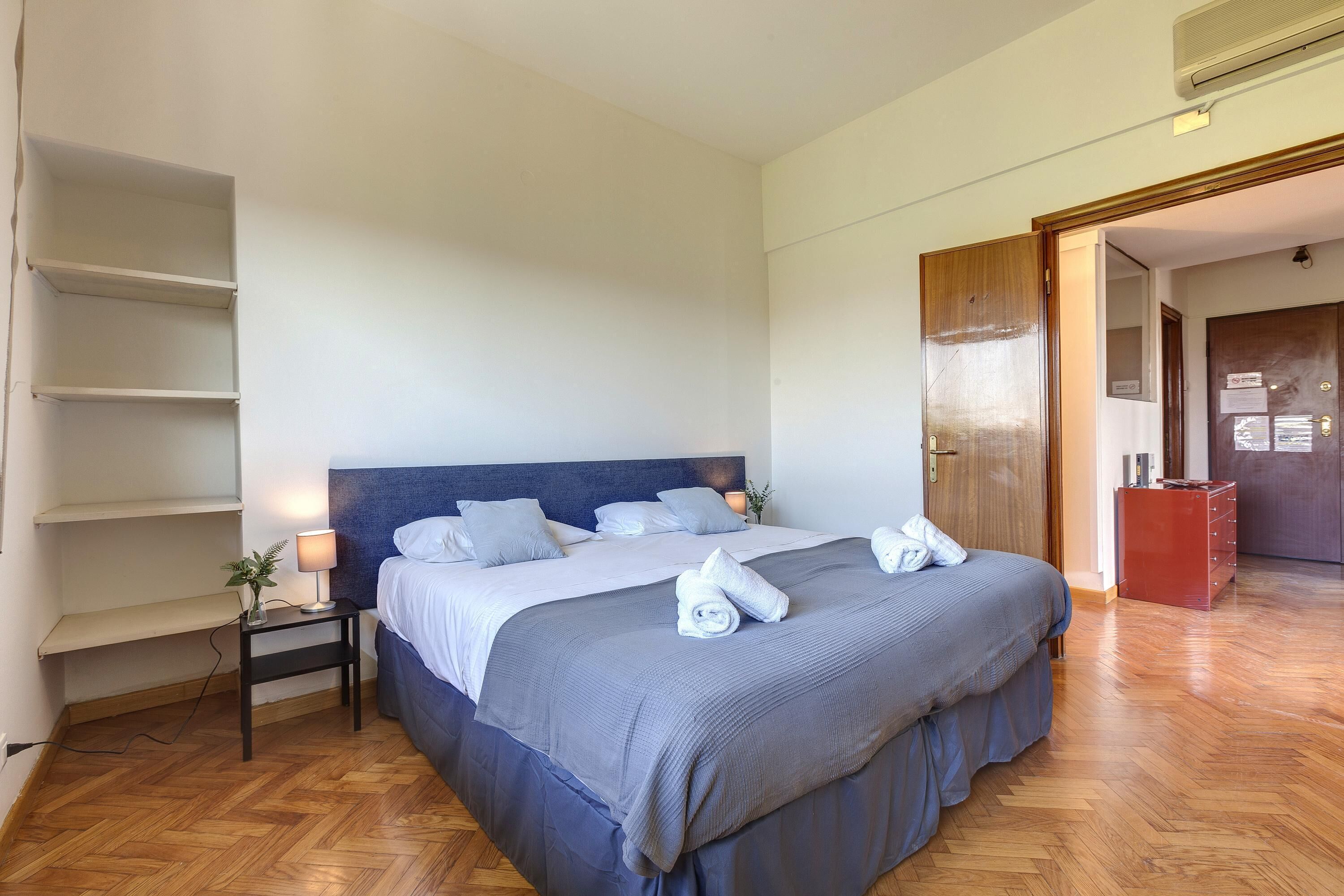 Uma cama bem arrumada com cabeceira azul, uma mesinha de cabeceira com abajur e uma cômoda vermelha em um quarto de hotel.