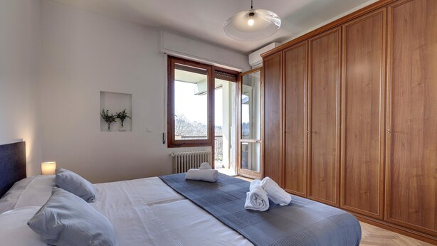 A neatly made bed with pillows, a bedside lamp, and a window with a view. - Colombo 76 in Firenze (Florence)