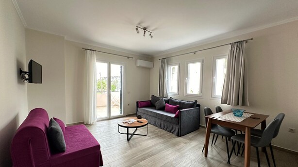 A living room with a purple sofa, a grey sofa, a wooden coffee table, and a dining table with chairs. There are large windows with curtains, a ceiling fan, and an air conditioning unit. - Michael Apartments - Private Parking (Lefkada)