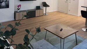 Un salon moderne avec un téléviseur à écran plat, un meuble en bois, un fauteuil noir, une table basse en verre et une porte.