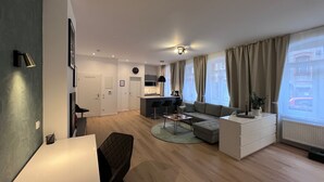 Un salon moderne comprenant un canapé, une table basse et un coin repas avec une table et des chaises. Il y a également un coin cuisine avec un évier et des placards. La pièce est dotée de grandes fenêtres avec des rideaux.