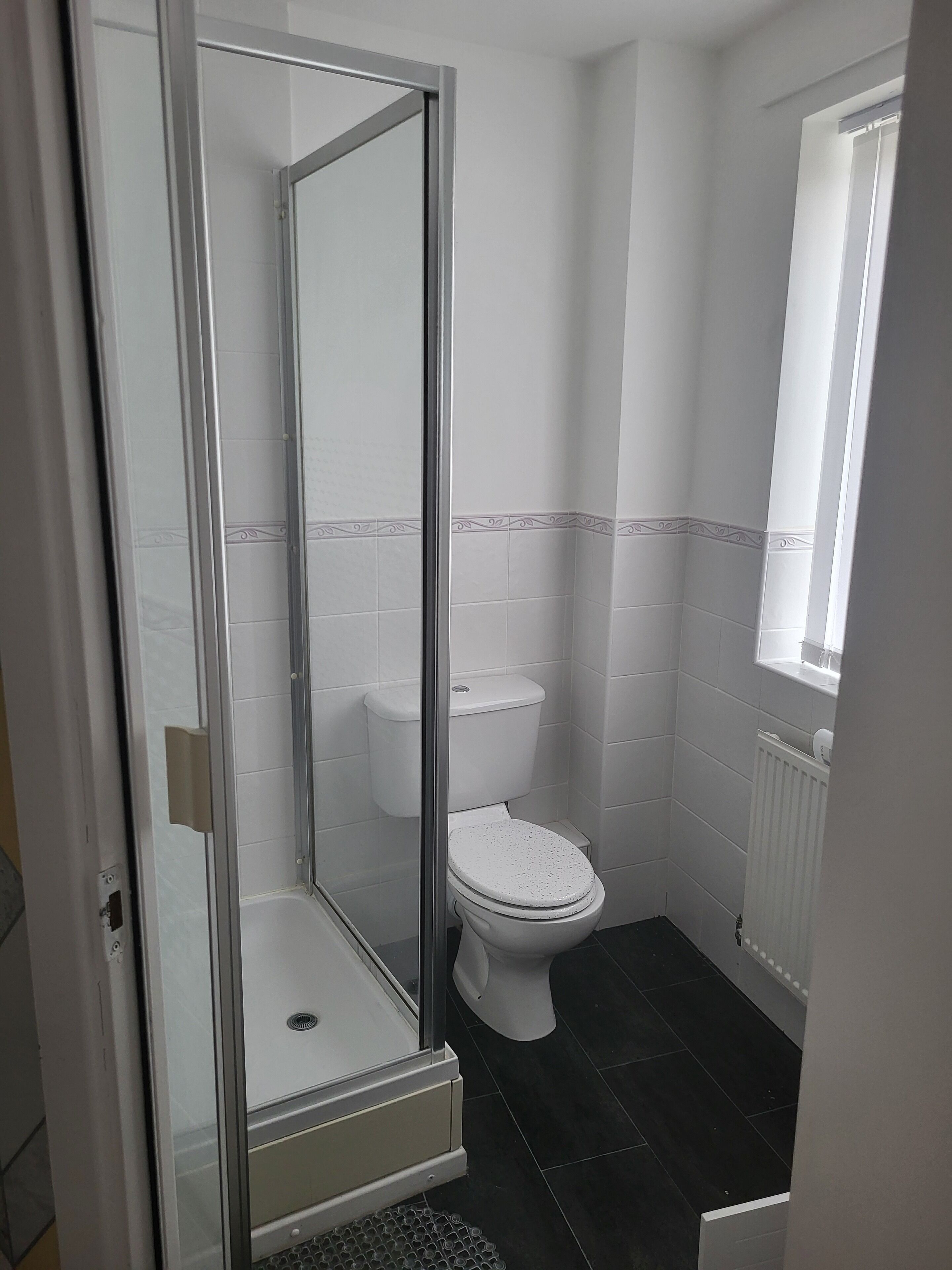A bathroom with a glass-enclosed shower, white tiles, a white toilet, and a window above the sink.