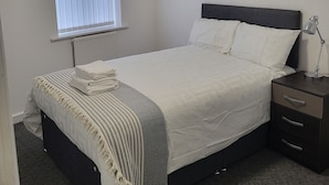 A neatly made bed with a white comforter and pillows, a bedside lamp, a dark nightstand with drawers, and a window with blinds. - Impeccable 4-bed House in Leicester (Leicester)