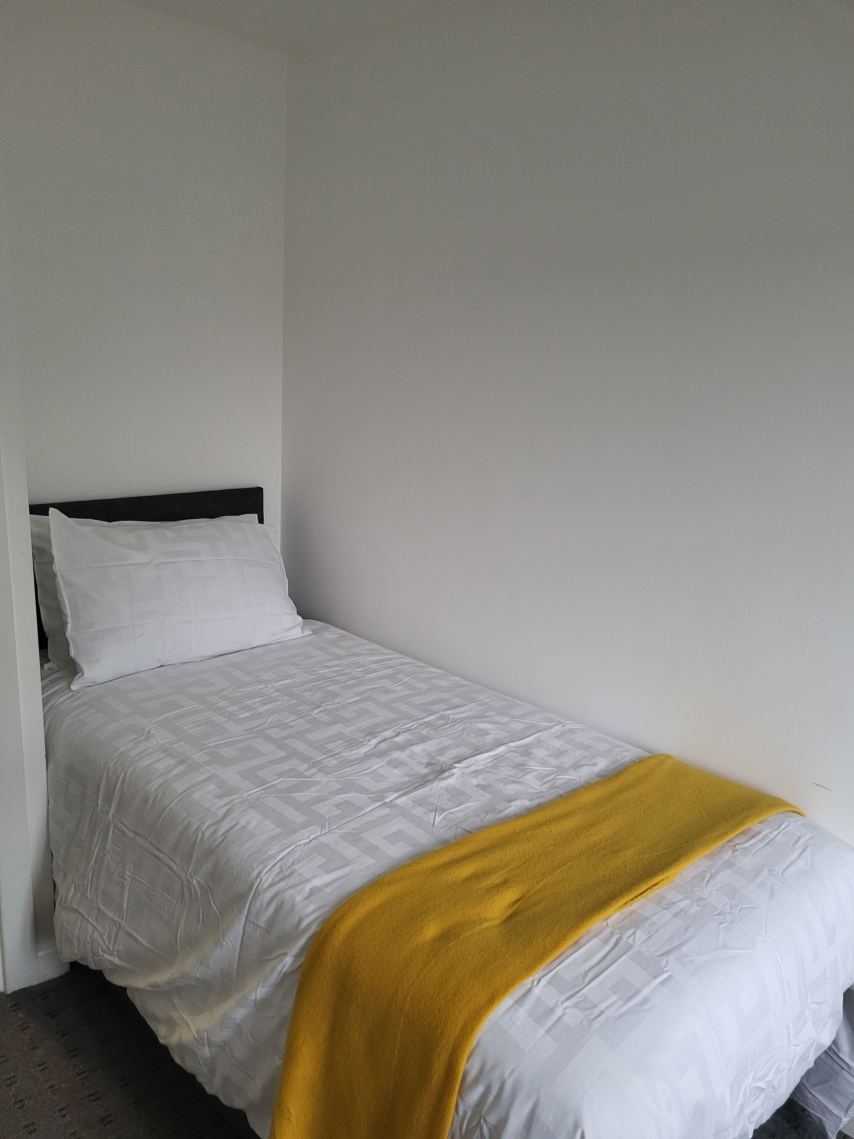 Un letto singolo con materasso bianco e coperta gialla, una testiera scura e una parete bianca liscia.