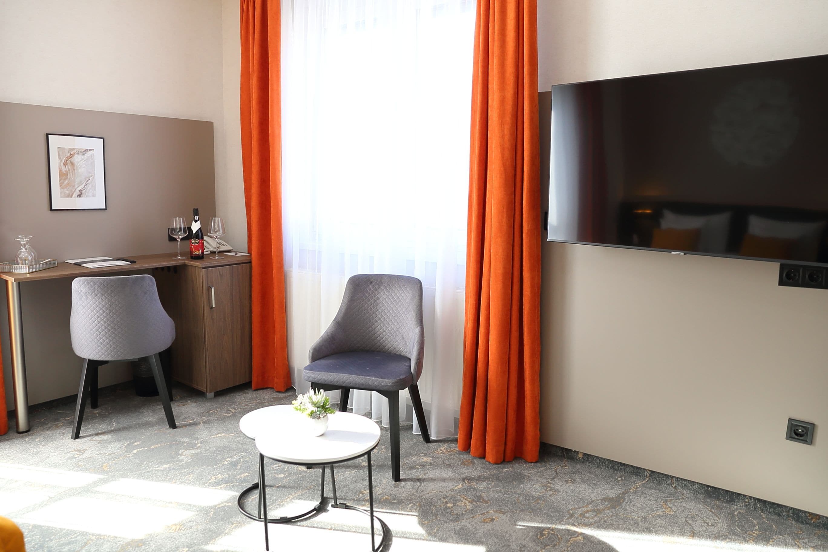 A hotel room with a desk, two chairs, a TV, and a window with curtains.