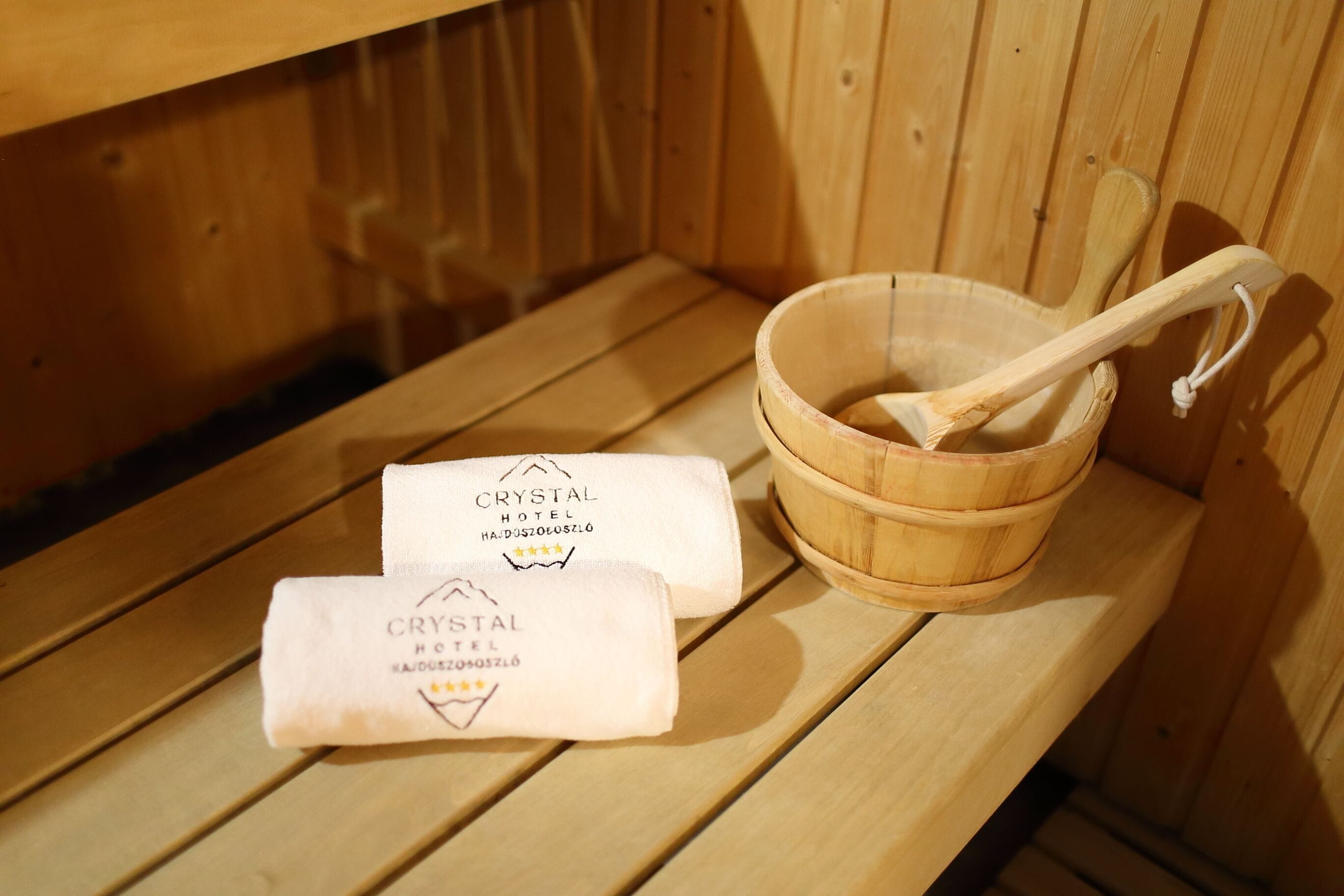 A wooden sauna bench with a wooden bucket and a wooden ladle. Two towels with the Crystal Hotel logo and name are placed on the bench.