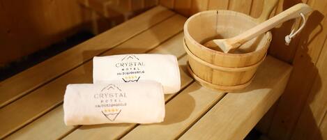 Eine Holzbänke in der Sauna mit einem Holzbecken und einem Holzlöffel. Zwei Handtücher mit dem Logo und dem Namen Crystal Hotel liegen auf der Bank.