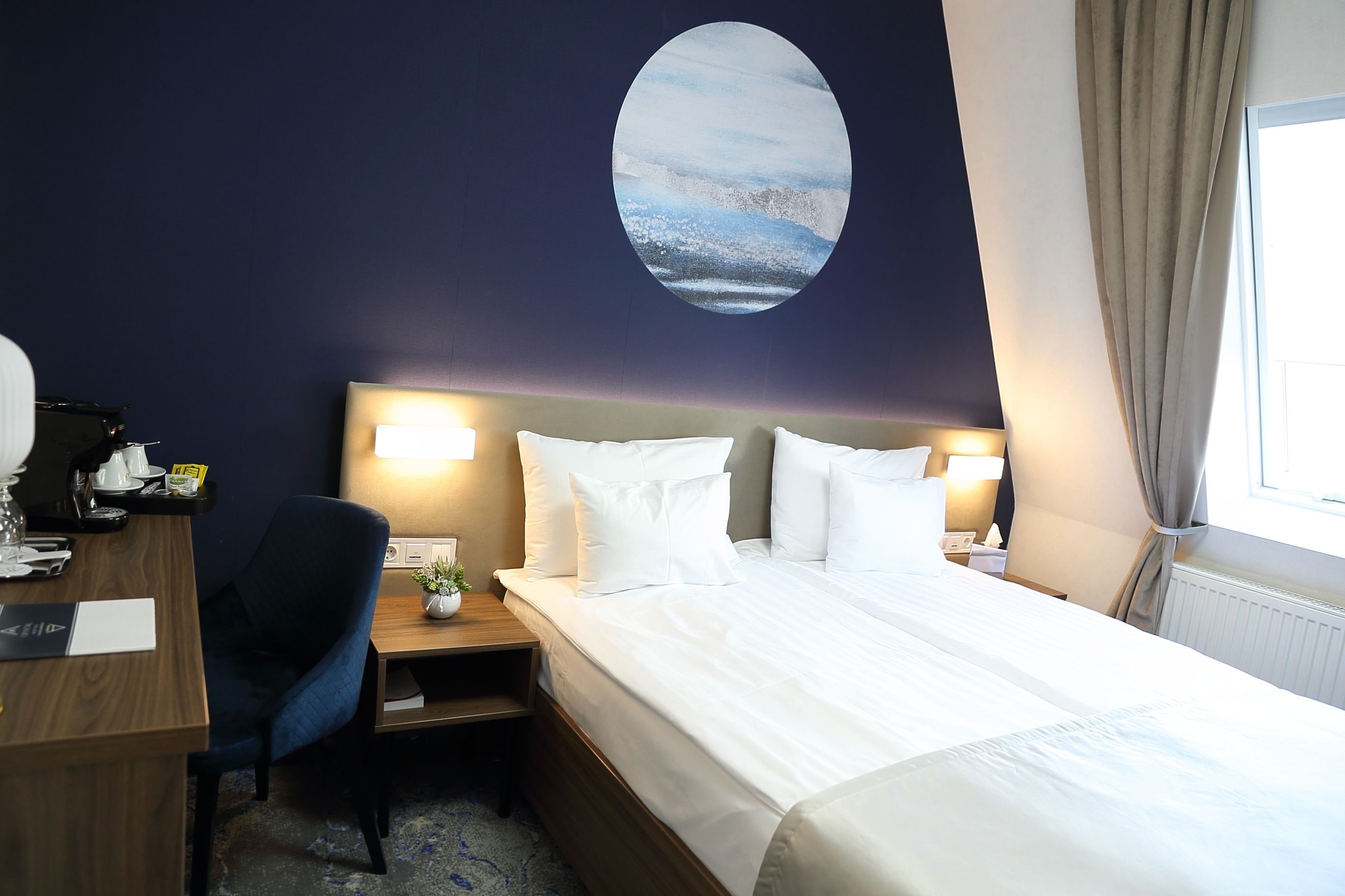 A hotel room with a bed, a desk, a chair, a nightstand, and a wall-mounted painting.