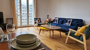Een moderne woonkamer met een blauwe bank, een houten salontafel en een eethoek met borden en een wijnglas.