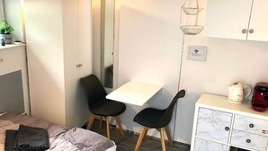 Una habitación pequeña con una cama, un escritorio, dos sillas, un armario blanco con cajones y una lámpara colgante.