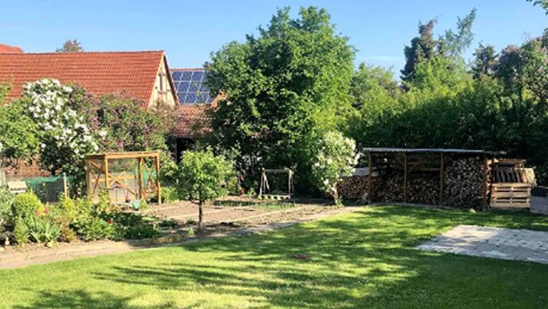Un jardín bien cuidado con césped, una estructura de juegos de madera, un jardín con diversas plantas y un edificio con techo rojo y paneles solares.
