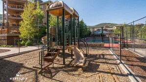 A playground with a slide and climbing structure, surrounded by a fence and a tennis court, with residential buildings in the background. - Alpine Abode at Canyons Village | Ski-In/Out Condo (Park City)