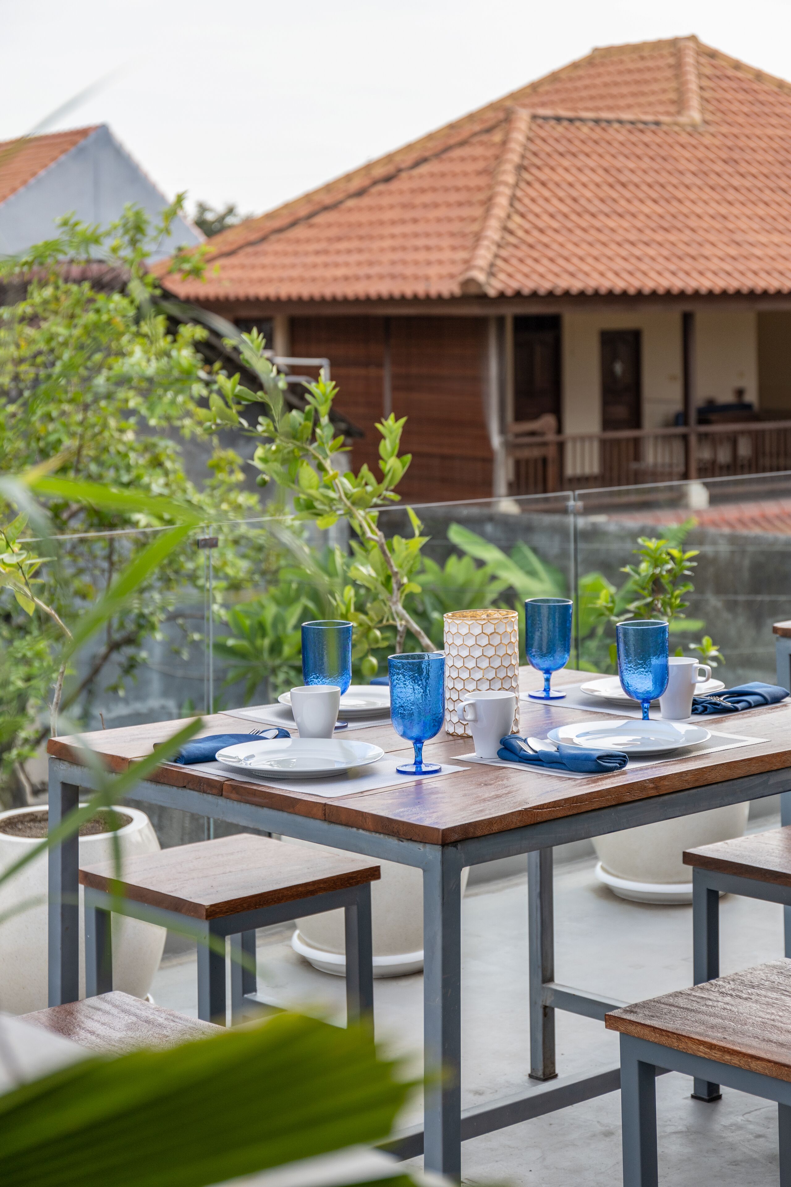 Une table à manger installée à l’extérieur, avec de la vaisselle en verre bleue et des assiettes blanches, entourée de plantes en pot et, au fond, d’un bâtiment dont le toit est recouvert de tuiles.