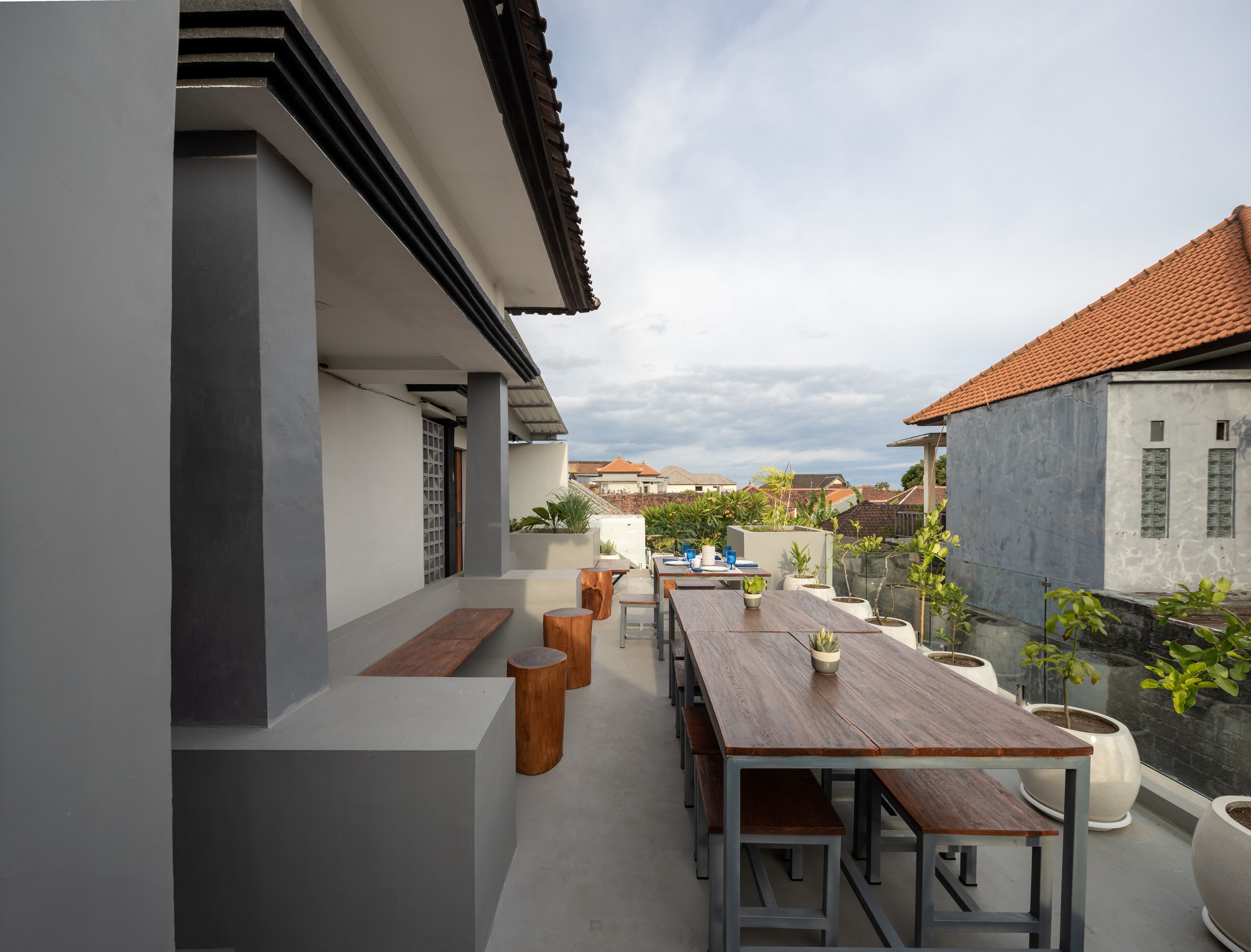 Una terraza en la azotea con una larga mesa de madera y bancos, rodeada de plantas en macetas y con vistas a una zona residencial.