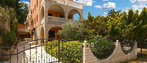 A multi-story building with a balcony, a gated entrance, and surrounding greenery.