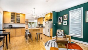 A modern kitchen with wooden cabinets, a central island with stools, and a dining area with a table and chairs. - Downtown Three Oaks Home, Sleeps 6, Fire Pit, Walk to Shops (Three Oaks)