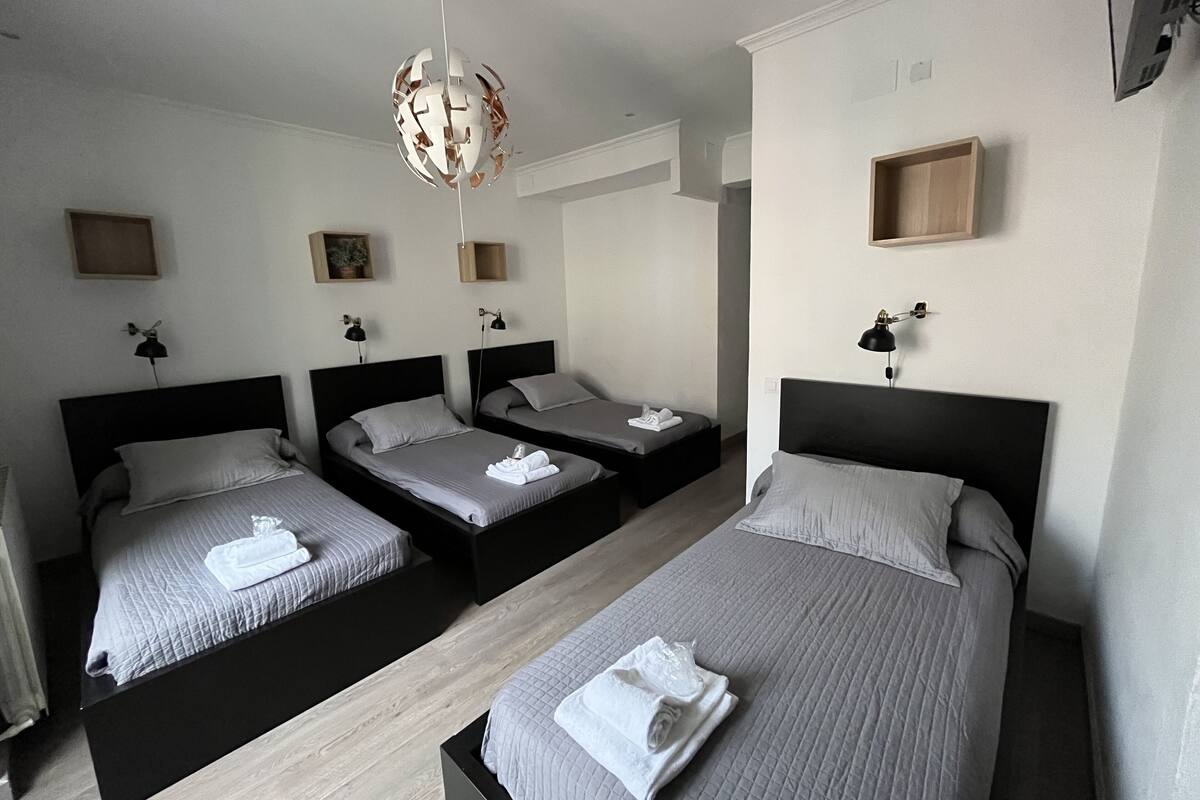 Habitación de hotel con tres camas, cada una con cubrecama gris y toallas blancas. Se observan mesitas de noche con lámparas, un candelabro moderno y un estante de madera con una planta.