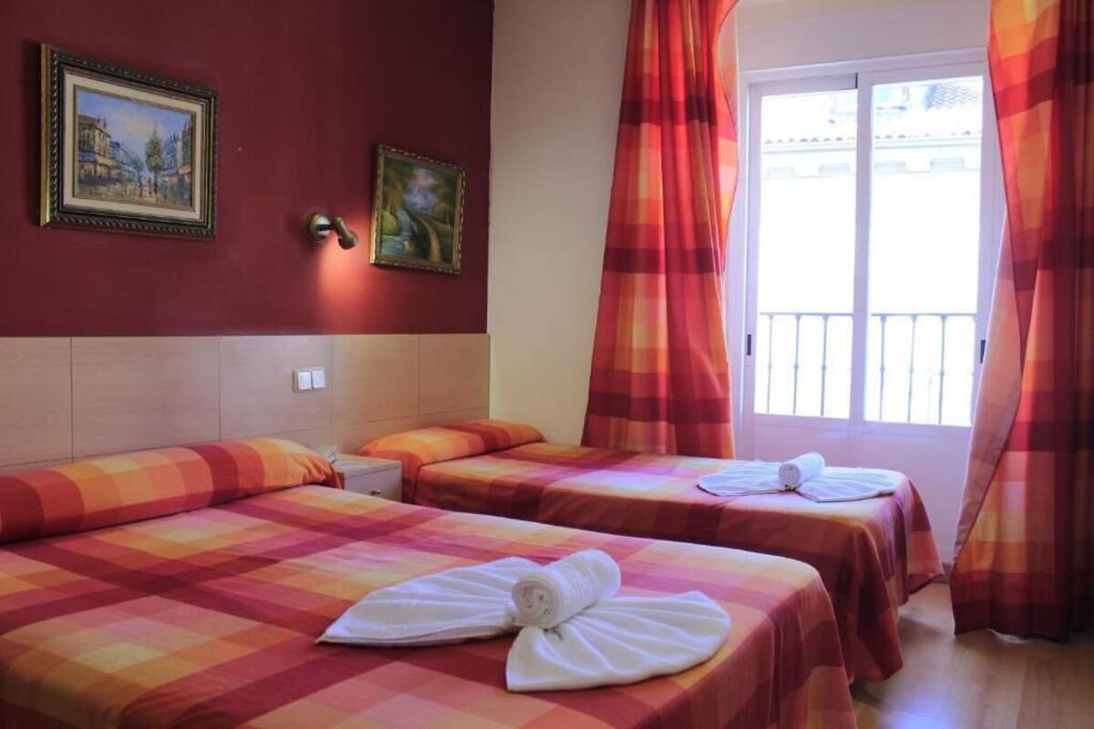 Habitación de hotel con dos camas, cada una con una colcha a cuadros y una toalla enrollada. Hay dos cuadros en la pared, y la habitación tiene una ventana con cortinas.