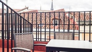 Une terrasse avec une table et deux chaises, une rambarde en métal et une vue sur des bâtiments aux toits recouverts de tuiles rouges et le clocher d’une église.