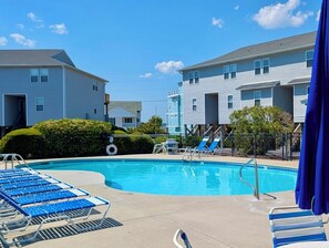 Une piscine avec des chaises longues bleues, un petit toboggan et un espace délimité par une clôture, sous un parasol bleu.