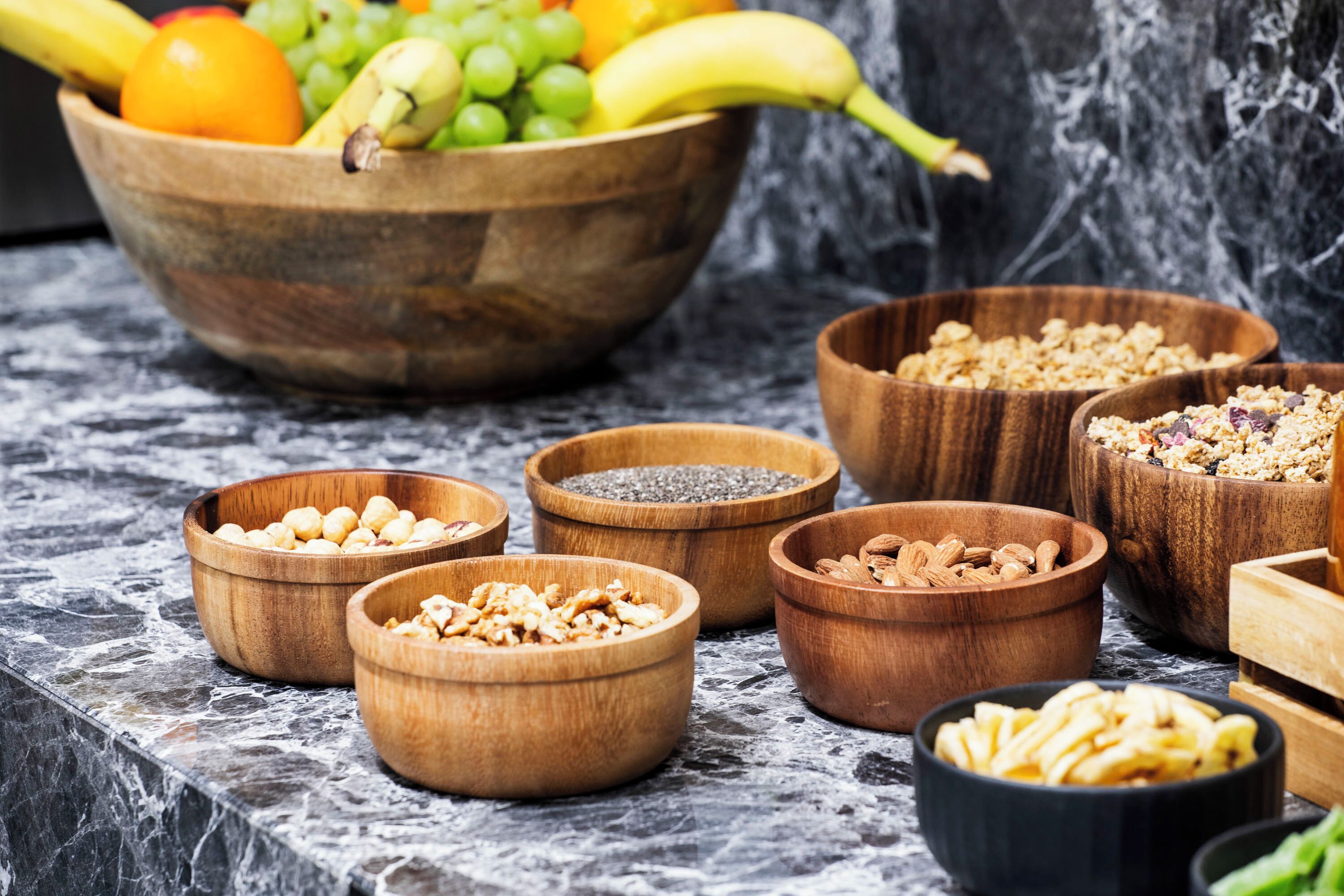A marble countertop with various wooden bowls containing fruits, nuts, and granola.