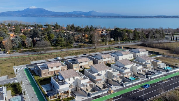 Un complexe résidentiel moderne composé d’immeubles compacts à plusieurs étages, doté d’une cour centrale et offrant une vue sur un lac et des montagnes en arrière-plan.