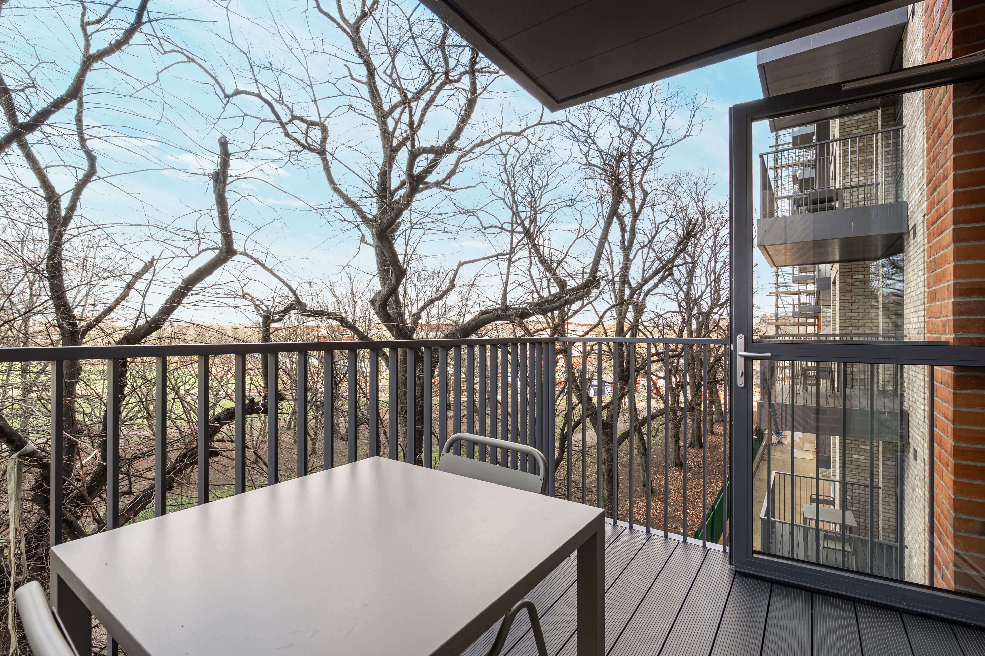 A balcony with a table and chairs, overlooking a brick building and bare trees.