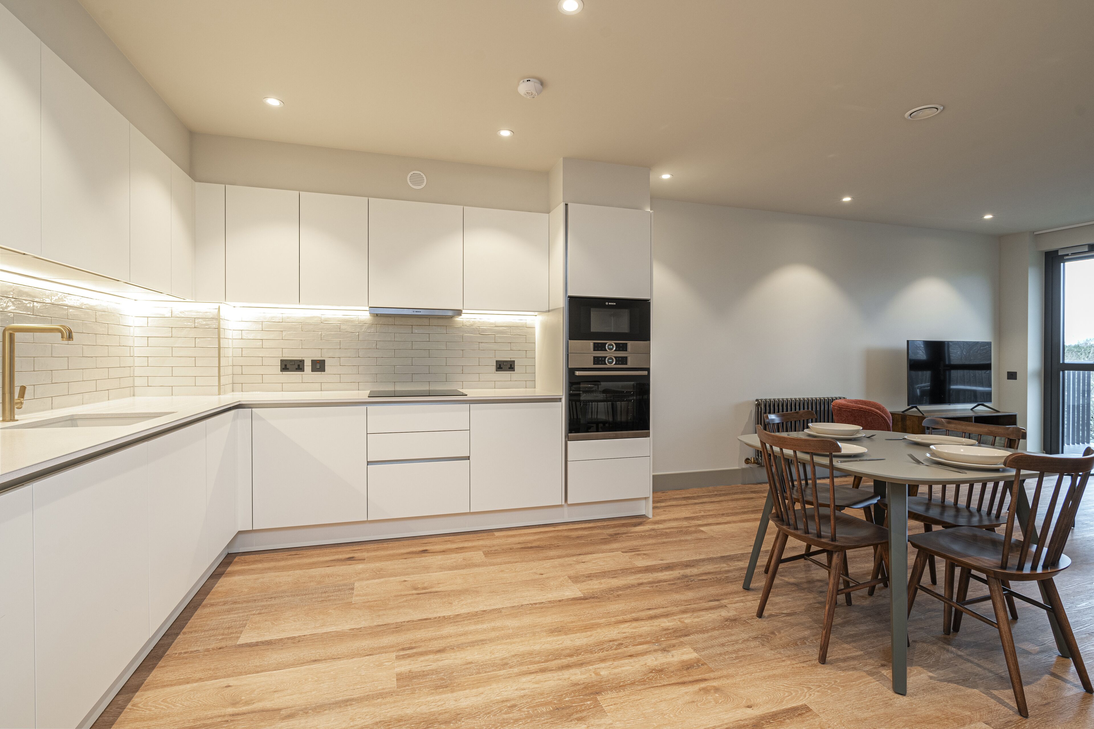 A modern kitchen with white cabinets, a built-in oven, and a dining area with wooden chairs and a table.