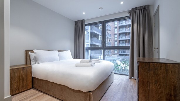 Habitación de hotel moderna con una cama grande, cabecera de madera y vistas a un paisaje urbano a través de ventanas de techo a suelo.