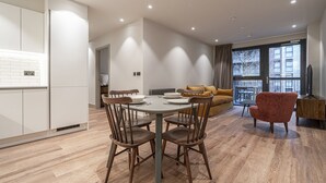 Un apartamento moderno con comedor, sala de estar con sofá y televisor, y cocina con gabinetes blancos y una ventana.