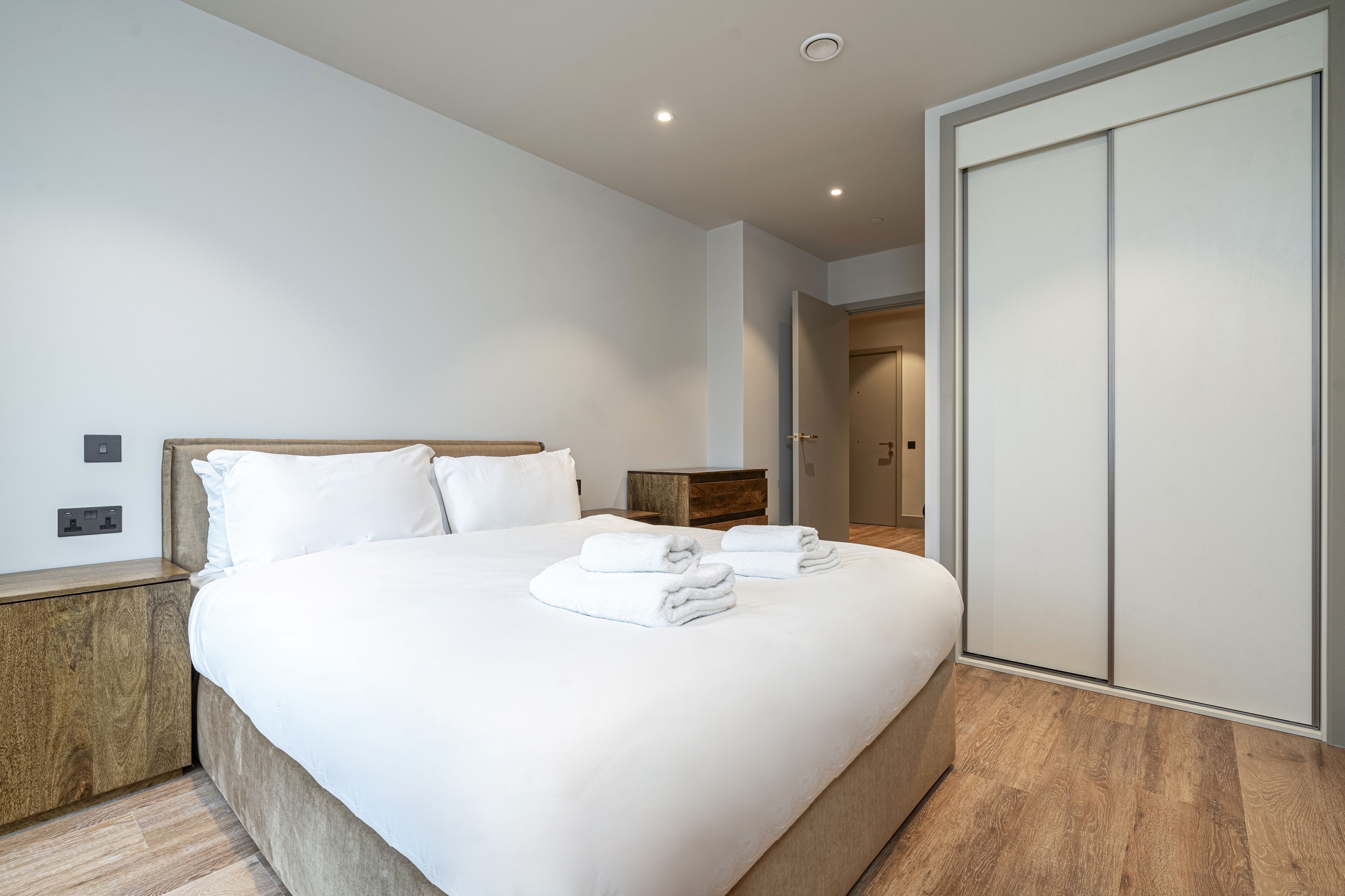 Ein modernes Hotelzimmer mit einem großen Bett, Holznachttischen, einem Kleiderschrank und einer neutralen Farbgestaltung.