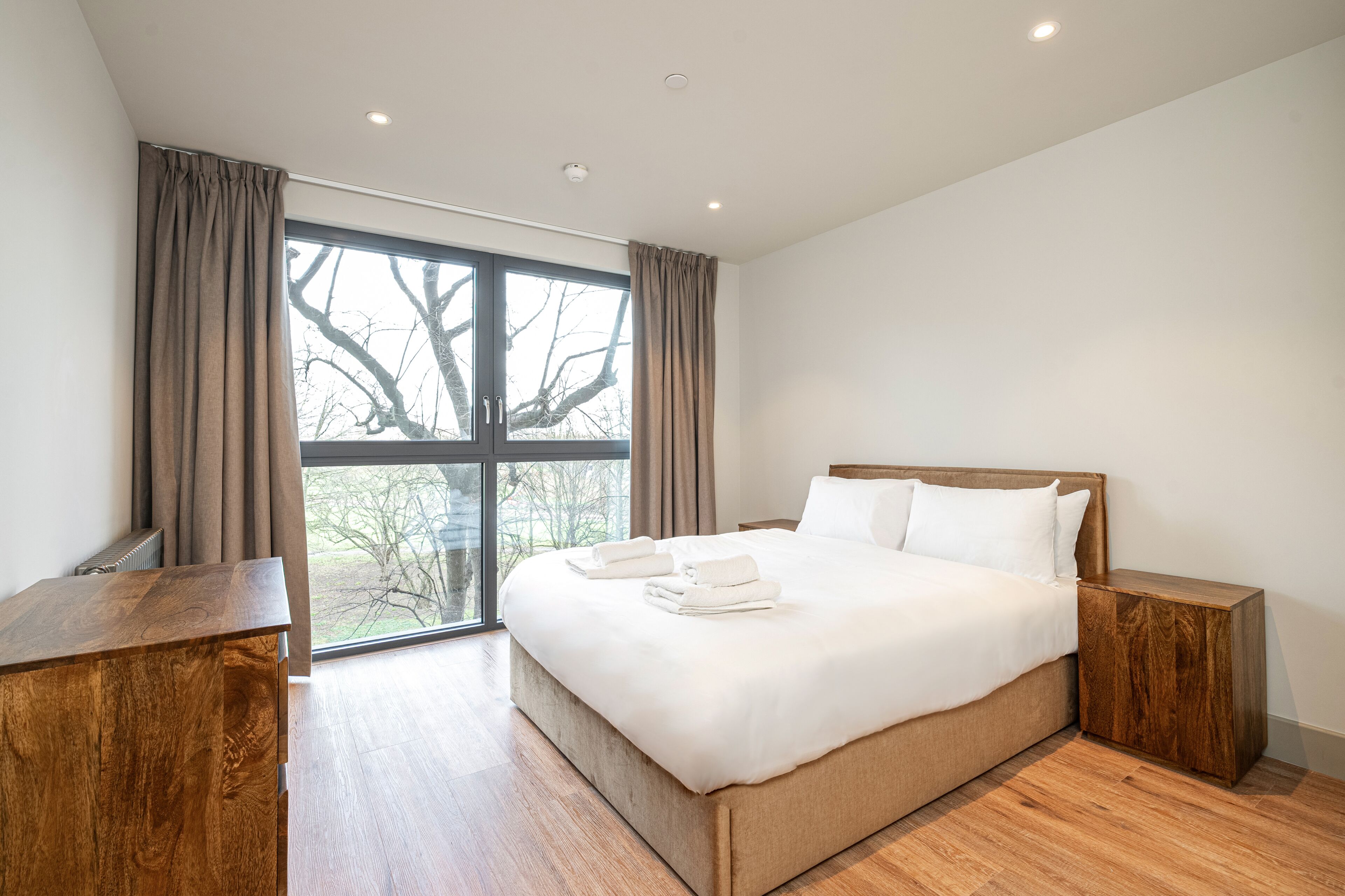 Habitación de hotel moderna con una cama grande, mesitas de noche de madera y vistas amplias por un ventanal.