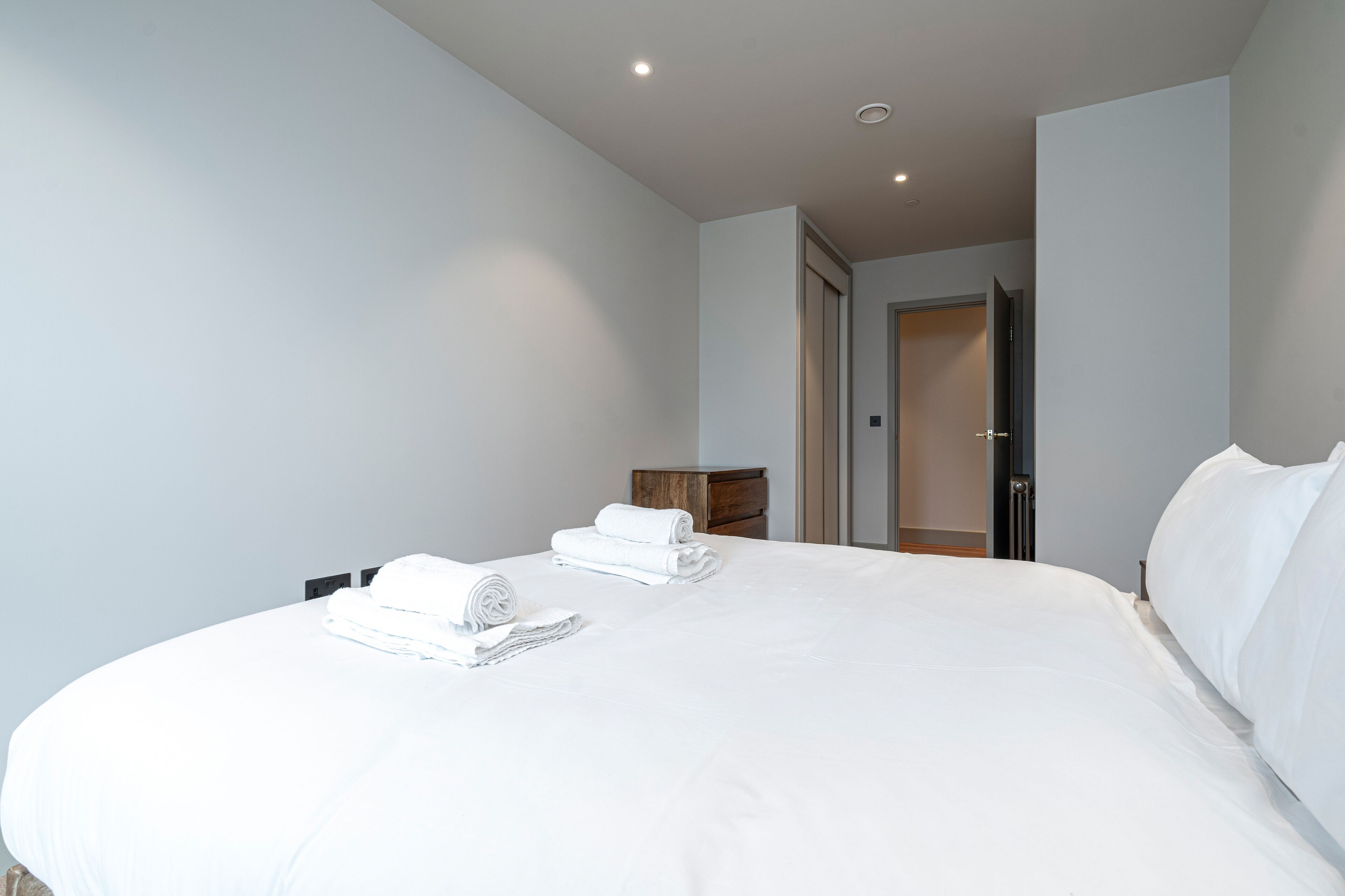 Een hotelkamer met een groot bed, twee net gevouwen handdoeken, een houten nachtkastje en een deur naar een andere kamer.