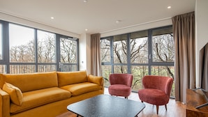 A modern living room with a mustard-colored sofa, two red armchairs, and a black coffee table. Large windows offer a view of trees and a building. - Spacious Flat in North London (London)
