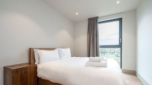 A modern hotel room with a large bed, a wooden nightstand, and a view of the city through a large window. - Spacious Flat in North London (London)
